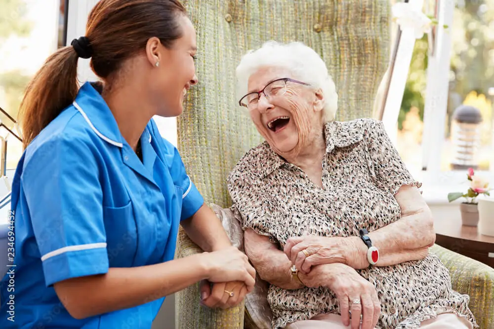 Senior Woman Sitting In Chair And Laughing With Nurse In Retirement Home